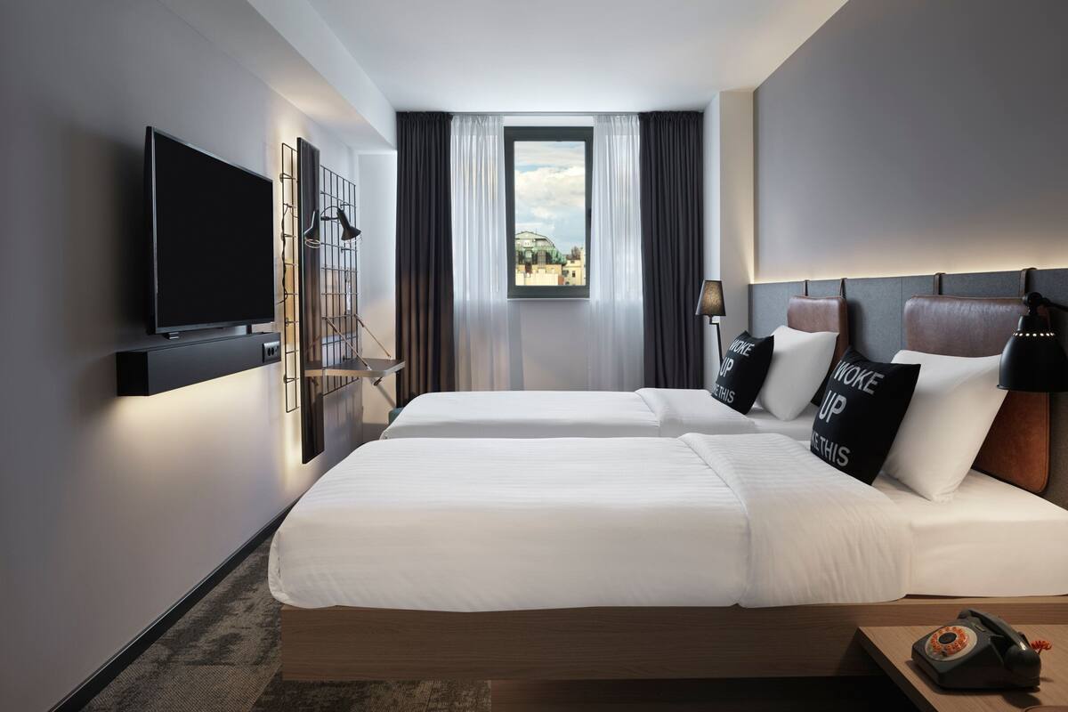 A hotel room with two beds, a TV mounted on the wall, a window with curtains, and a lamp on the bedside table.