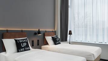 A hotel room with two beds, each with a pillow that says "I woke up like this." There is a lamp on the right side of the room and a window with curtains.