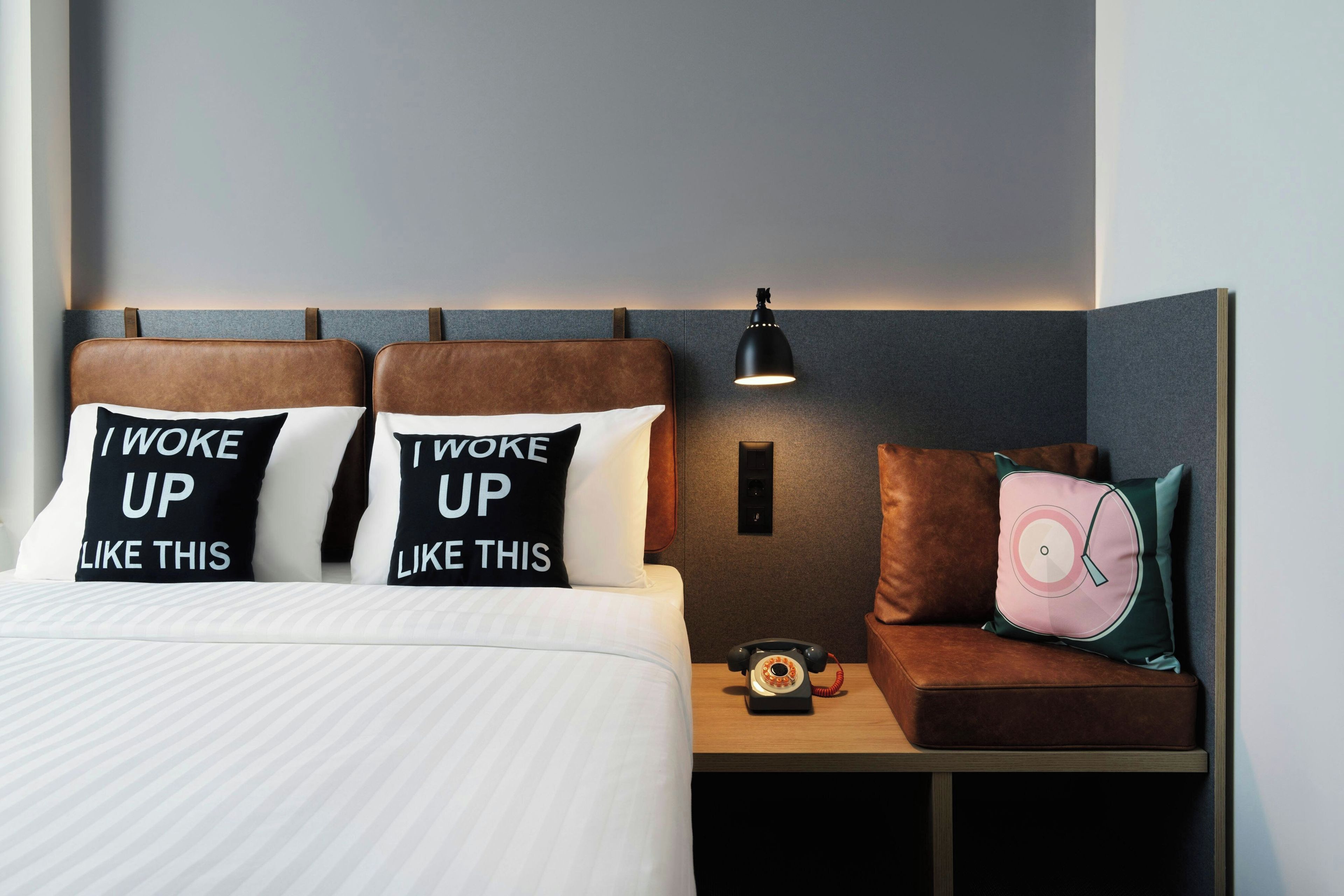 A hotel room with a bed, a nightstand with a lamp, and two pillows with motivational text.