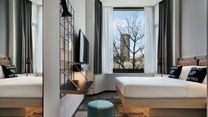 A modern hotel room with two beds, a large window with a view of a building and trees, and a small seating area with a table and chairs. - Moxy Belgrade (Belgrade)
