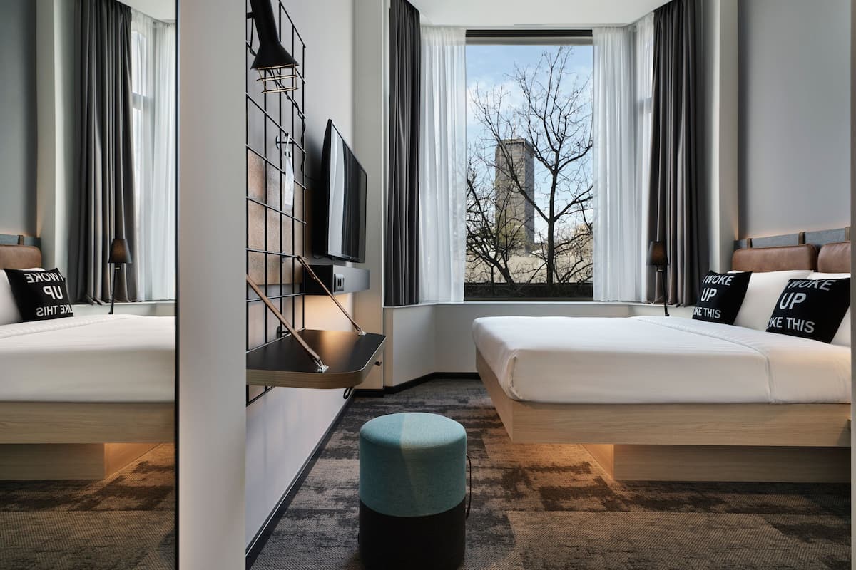 A modern hotel room with two beds, a large window with a view of a building and trees, and a small seating area with a table and chairs.
