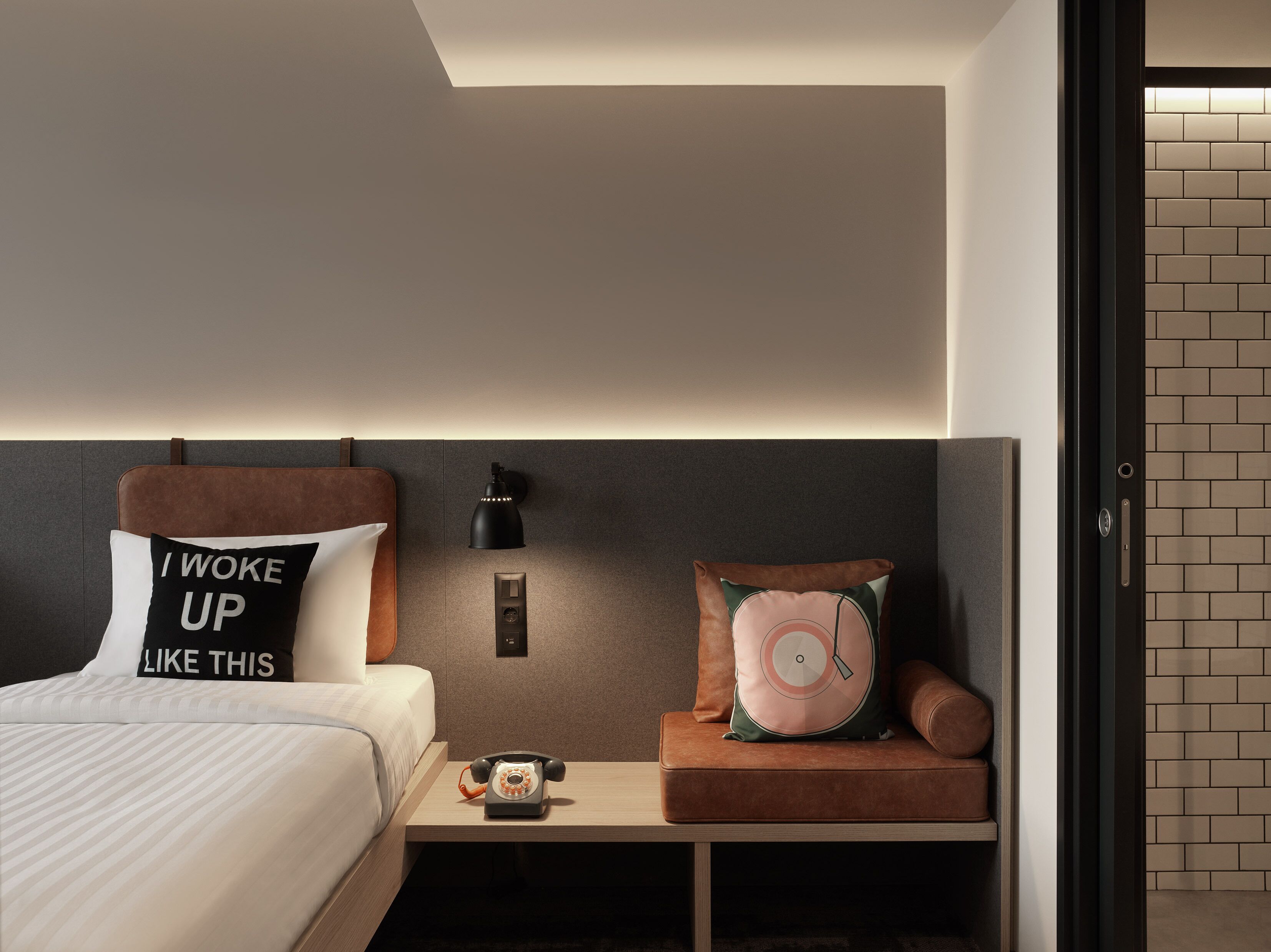 A modern hotel room with a neatly made bed, a bedside table with a vintage telephone, a wall-mounted lamp, and a small seating area with a cushioned bench.