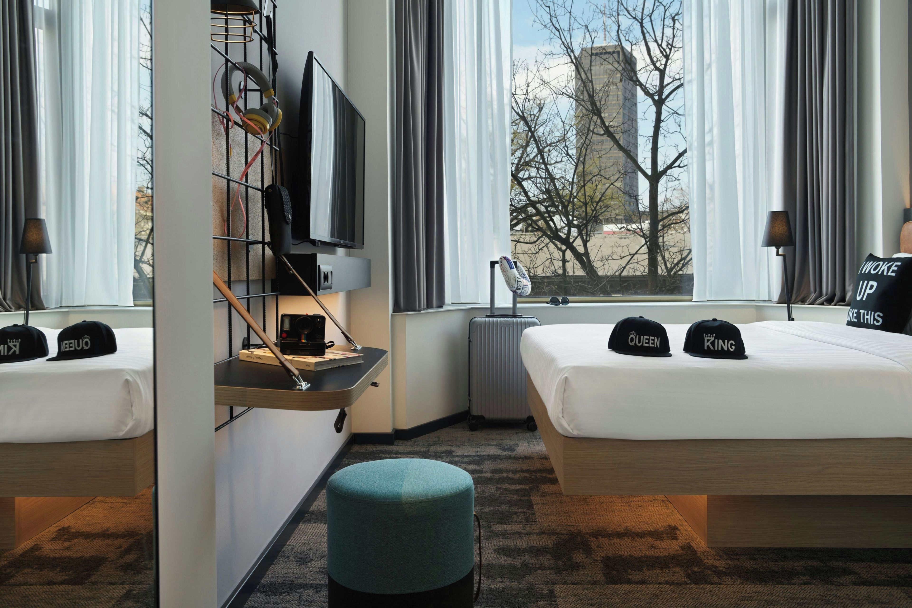 Habitación de hotel con dos camas, un escritorio con computadora, televisión y vista a un edificio y árboles.