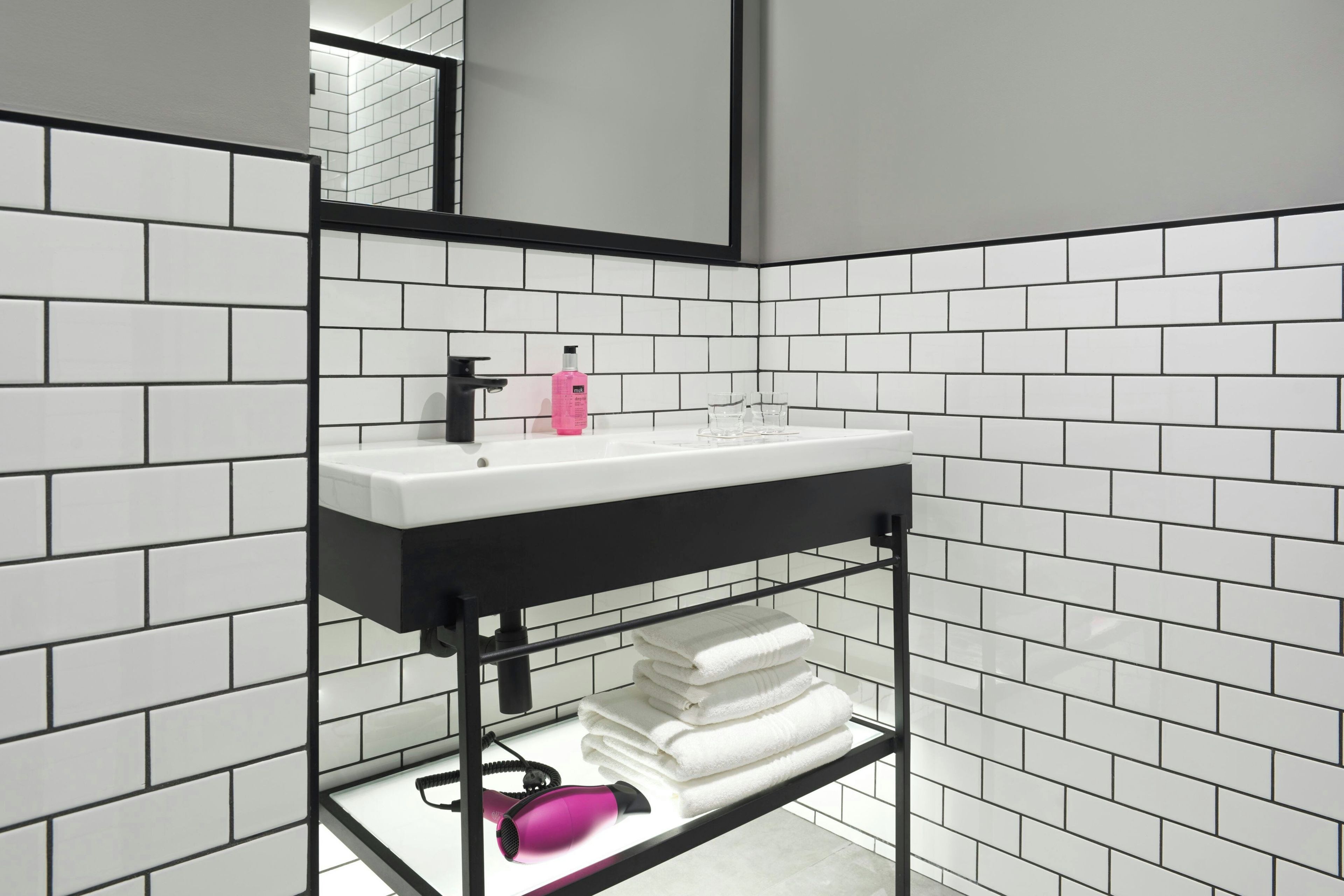 Un baño con un lavamanos blanco, un grifo negro y un espejo encima. Hay azulejos blancos en las paredes y un estante con toallas y un secador de pelo.