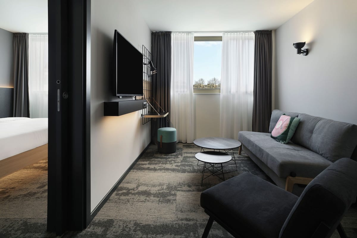 A modern hotel room with a bed, a grey sofa, a small round table, and a TV mounted on the wall.
