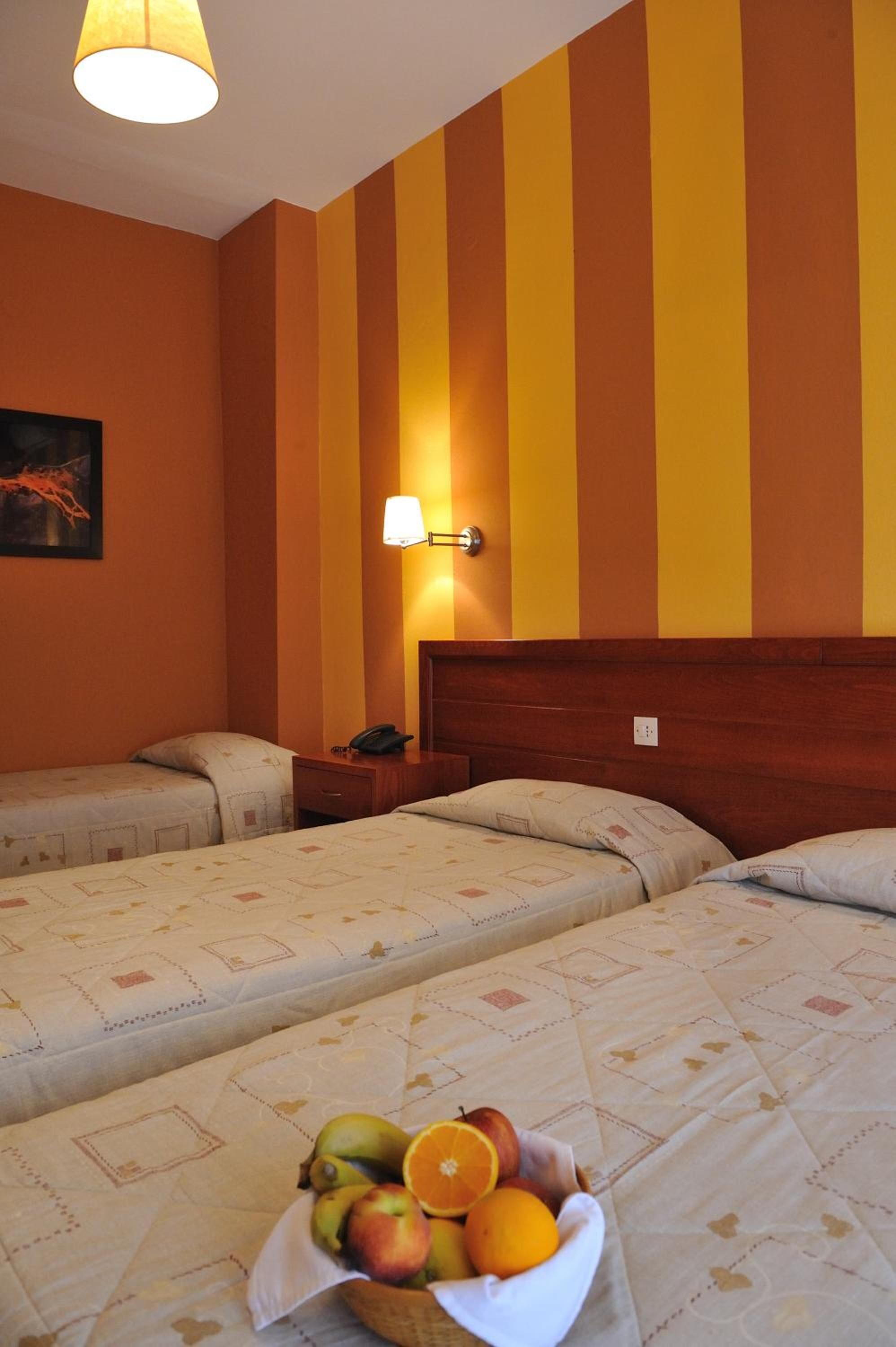 A hotel room with two beds, a nightstand, a lamp, and a fruit basket on the bed.