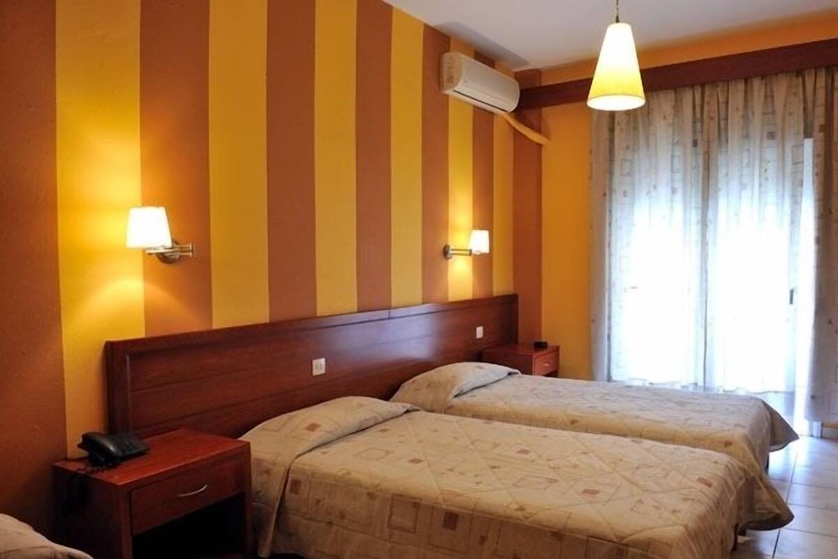 Une chambre d’hôtel avec deux lits, des têtes de lit en bois, des tables de chevet avec des lampes et une grande fenêtre avec des rideaux.