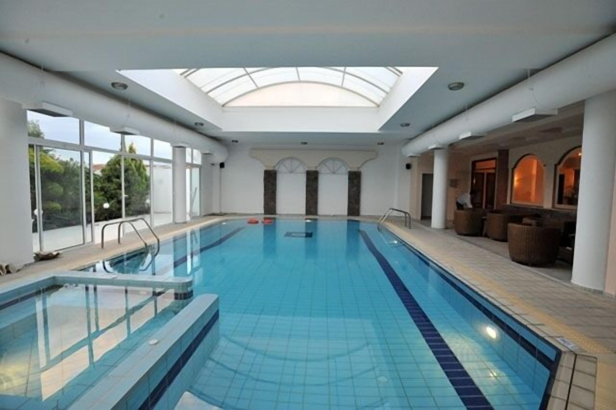 Una piscina interior con tragaluces, paredes de vidrio transparente y un área de descanso con asientos.