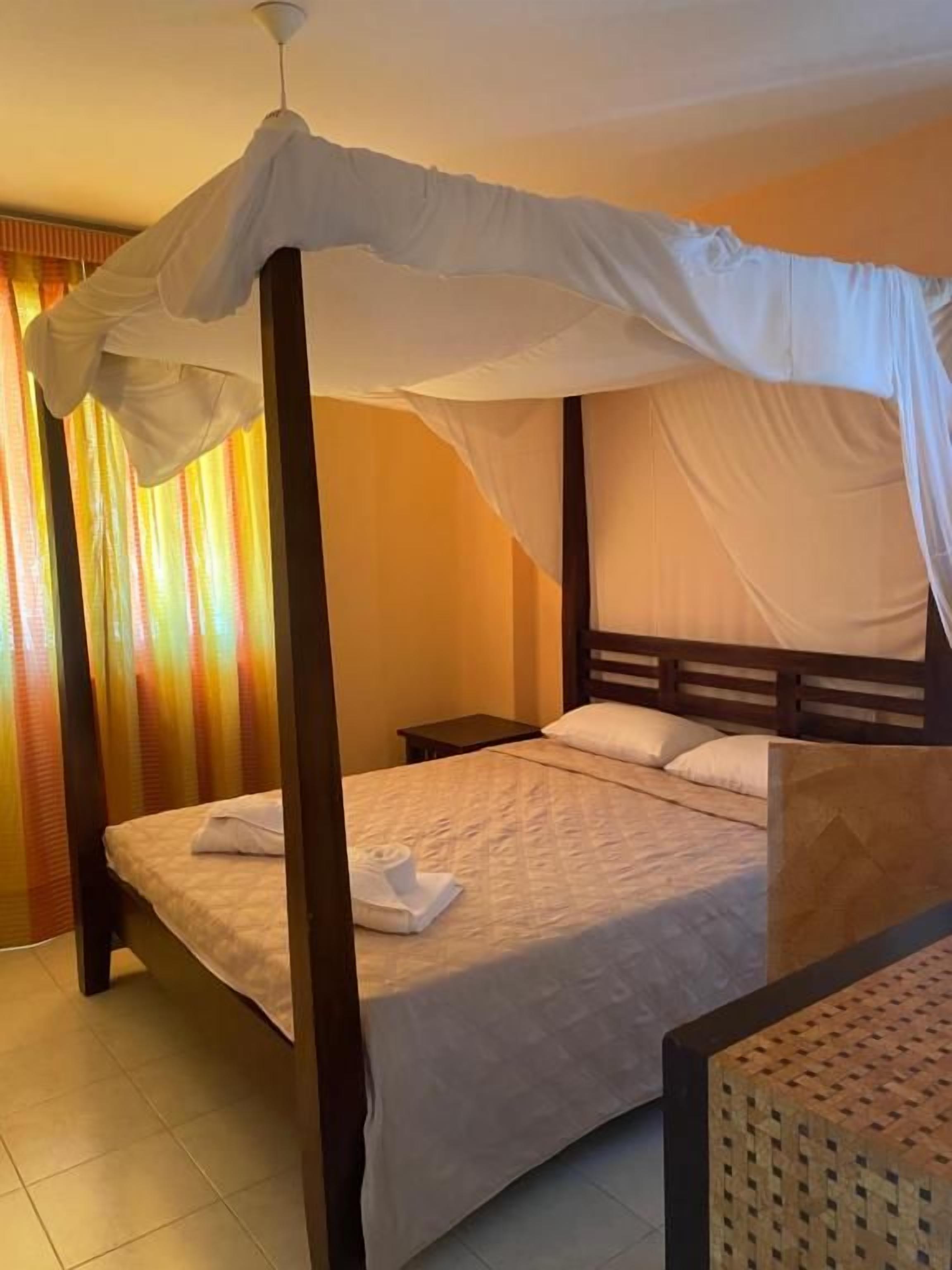 Una cama con dosel y pilares, cabecera de madera y mesita de noche con lámpara.
