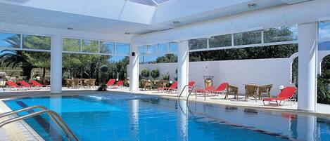 An indoor swimming pool with clear blue water, surrounded by red and white lounge chairs, and large windows allowing natural light.