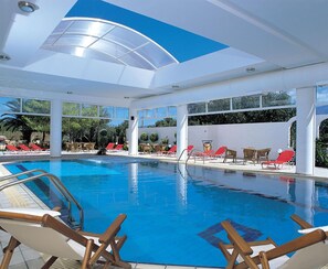 Una piscina interior amb aigua blava cristal·lina, envoltada de gandules vermelles i blanques, i grans finestres que deixen entrar llum natural.