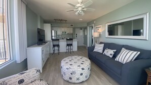 Una sala de estar moderna con sofá, reposapiés y sillones, una cocina al fondo y un ventilador de techo.