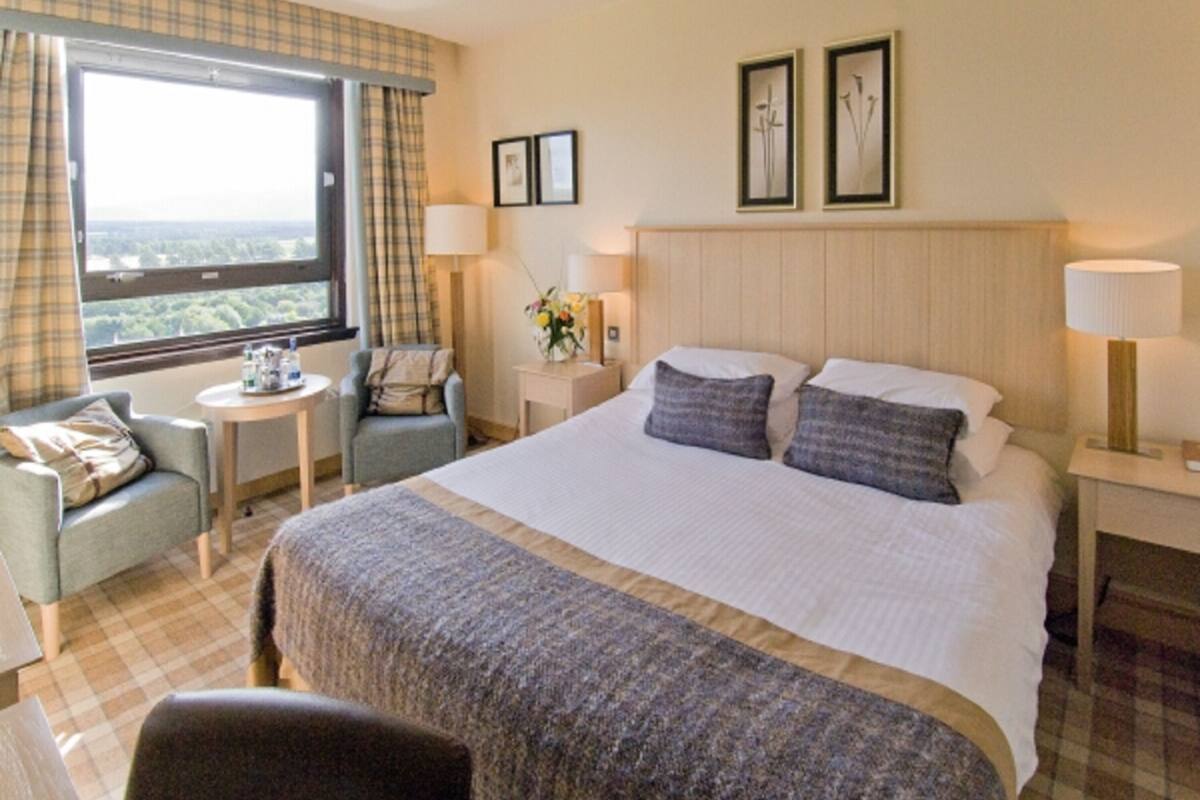 A hotel room with a large bed, a small armchair, a round table, and a window with a view.