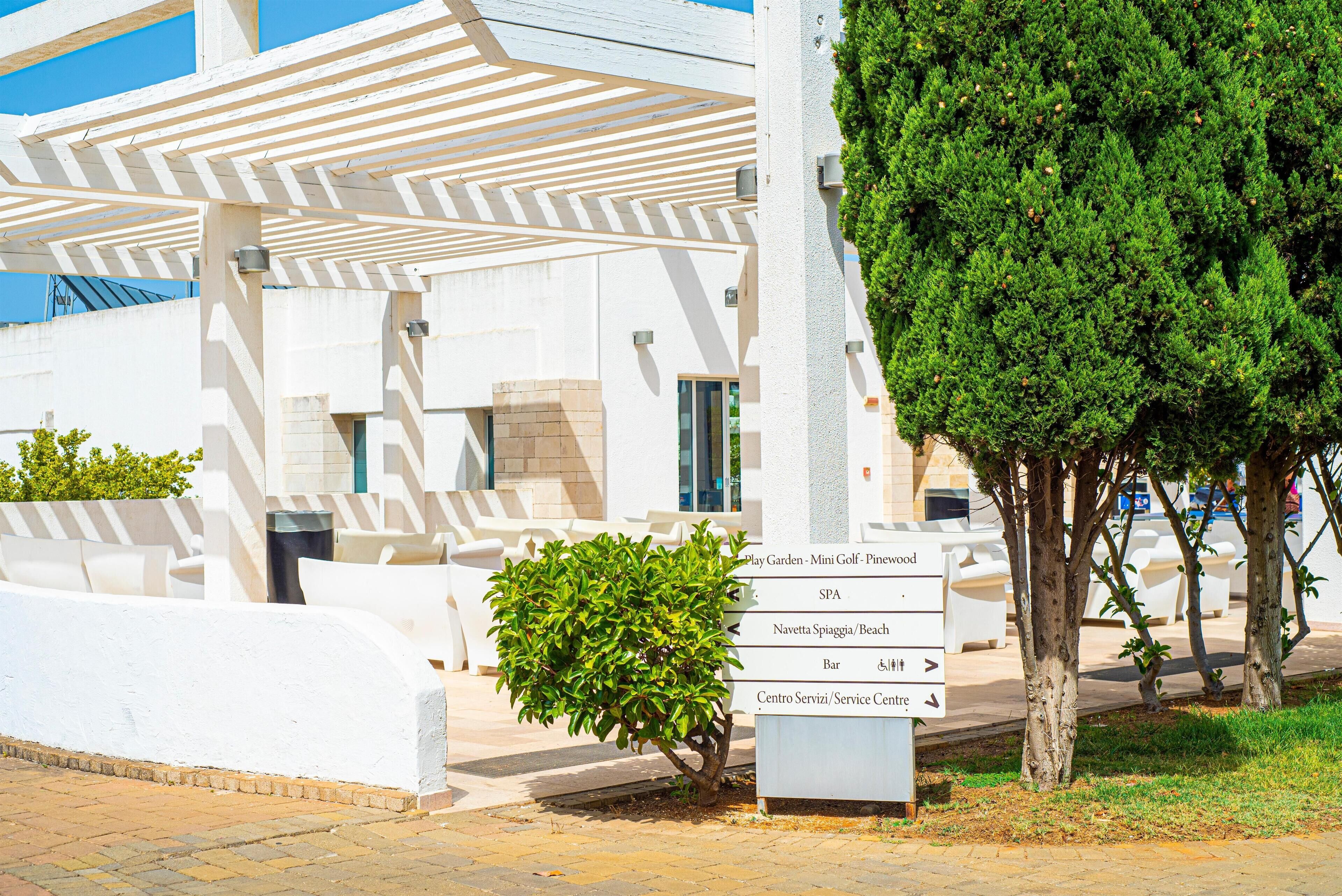A building with a white exterior, a covered outdoor seating area, and a sign indicating amenities including a spa, beach, and service center.