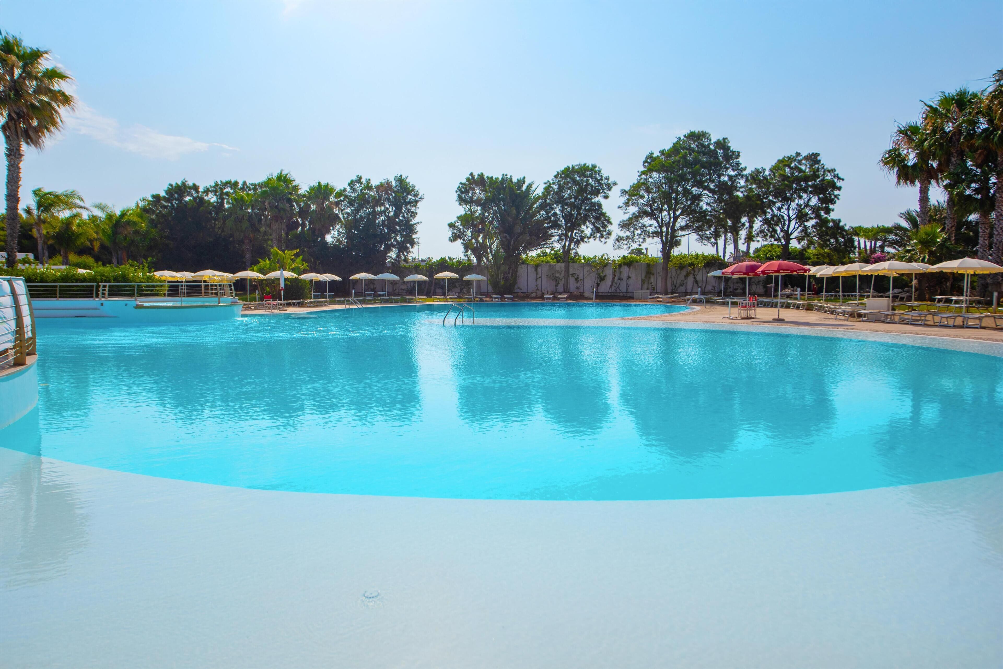 A large swimming pool with a clear blue water, surrounded by palm trees and shaded by umbrellas.