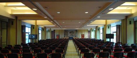 Un salón de conferencias con filas de sillas, piso alfombrado y una pantalla grande al frente.