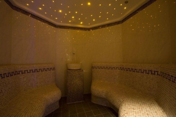 A room with two mosaic-tiled benches, a small sink, and a ceiling with recessed lighting.