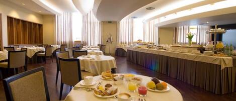 Espaço de refeições de hotel com mesas redondas, cadeiras e um buffet.