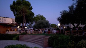 Evento al aire libre con mesas y sillas dispuestas en un área pavimentada, rodeado de árboles y un edificio al fondo.