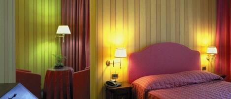 Habitación de hotel con una cama, dos mesitas de noche con lámparas, un escritorio con una silla y una pequeña mesa con una planta.