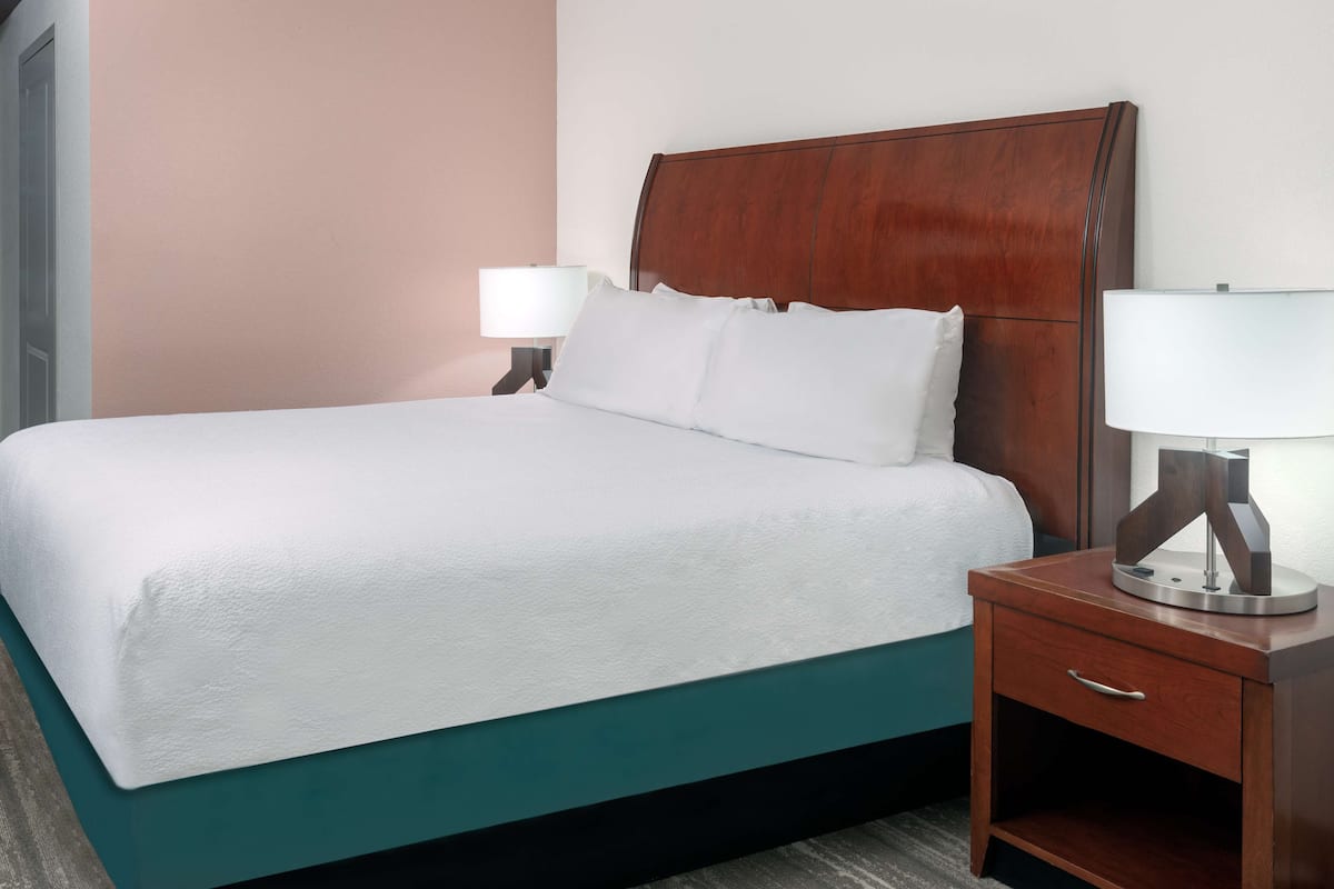 Una cama bien hecha con sábanas blancas, cabecera de madera y mesita de noche con lámpara.