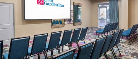 Une salle de conférence avec des rangées de chaises bleues, un grand écran affichant le logo du Hilton Garden Inn et un sol recouvert de moquette.