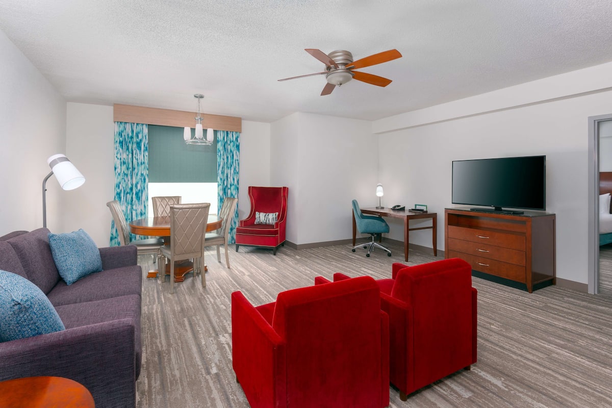 Habitación de hotel con sofá, sillas rojas, escritorio, televisor y ventilador de techo.