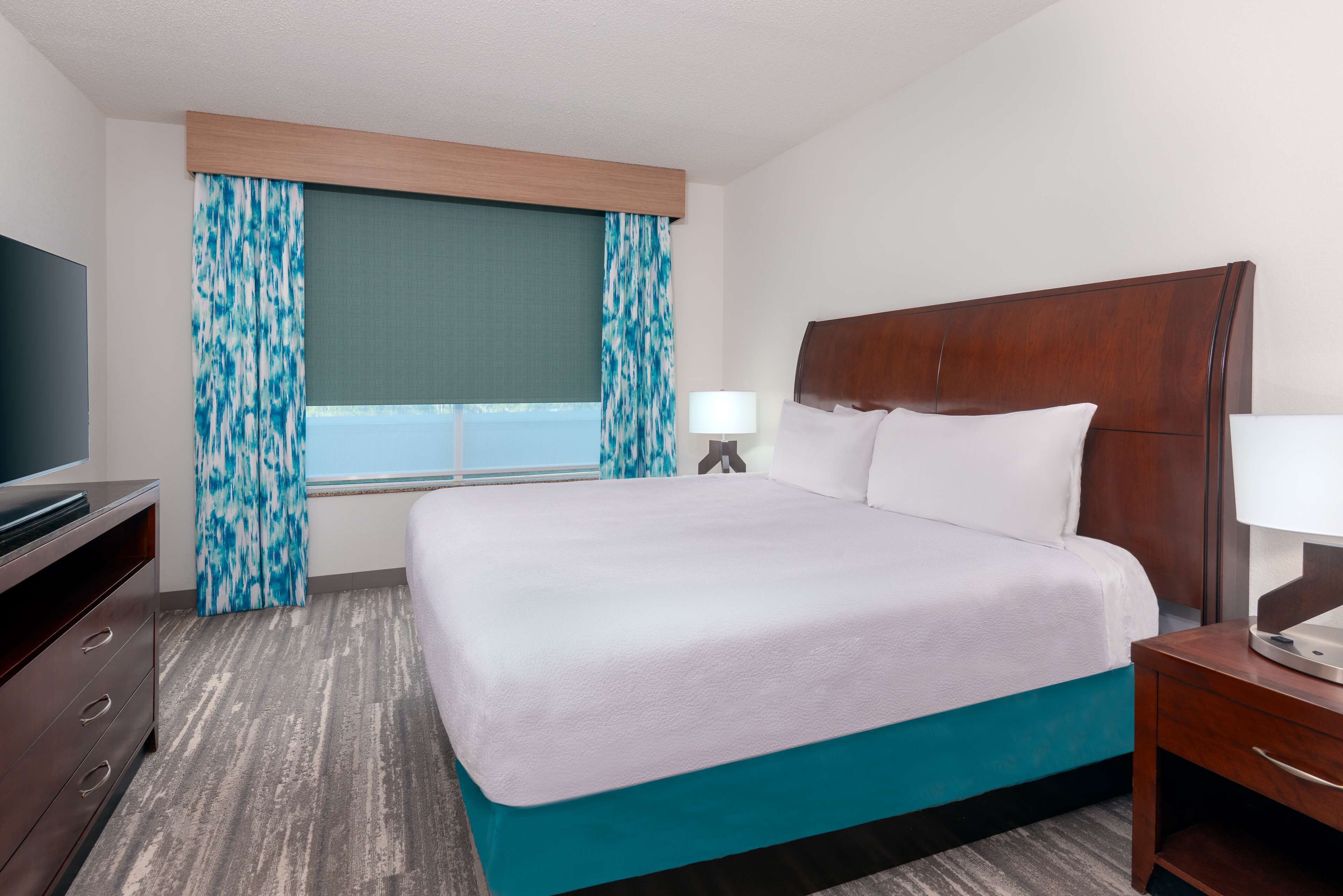 Ein Hotelzimmer mit einem großen Bett, einem Fernseher auf einer Kommode und einem Fenster mit Vorhängen.