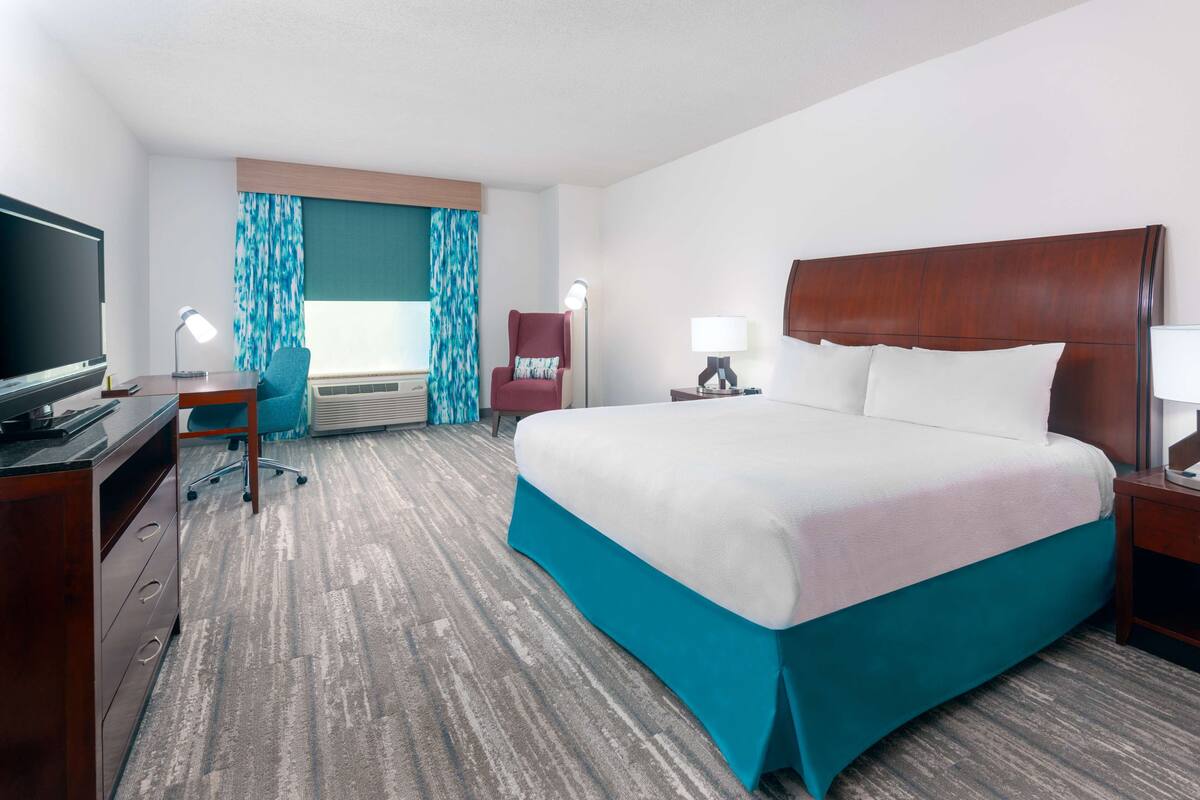Habitación de hotel con una cama grande, un escritorio con televisor, una silla, una lámpara y un ventanal con cortinas.