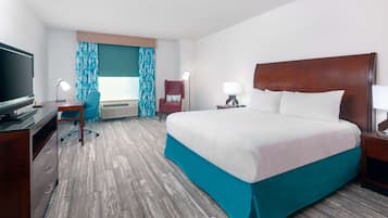 A hotel room with a large bed, a desk with a TV, a chair, a lamp, and a window with curtains.