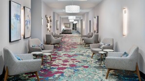 Ein Hotelkorridor mit Teppichmuster, grau gepolsterten Sesseln und einem zentralen Kronleuchter.
