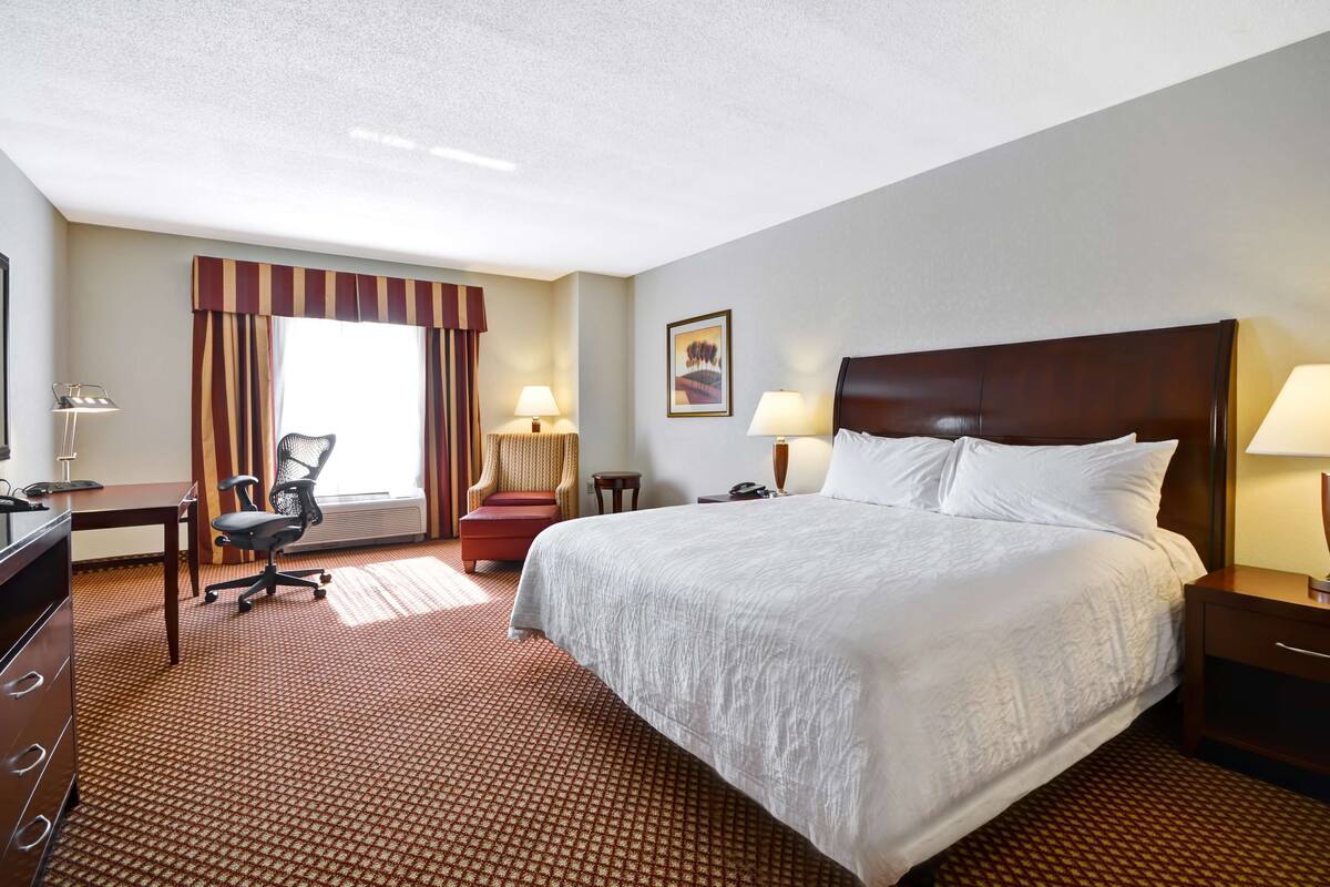 Una habitación de hotel con una cama grande, un escritorio, una silla, una lámpara y un ventanal con cortinas.