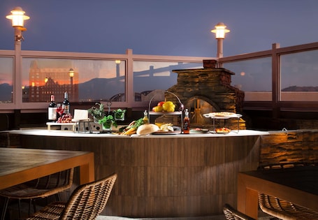 Zona de bar al aire libre con horno de piedra, sillas de mimbre y vistas a un paisaje urbano al anochecer.