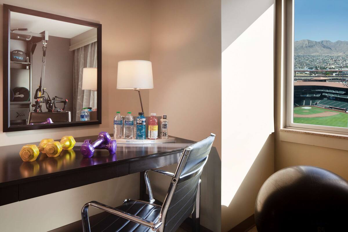 A hotel room with a desk, a chair, a mirror, a lamp, and a view of a baseball stadium.