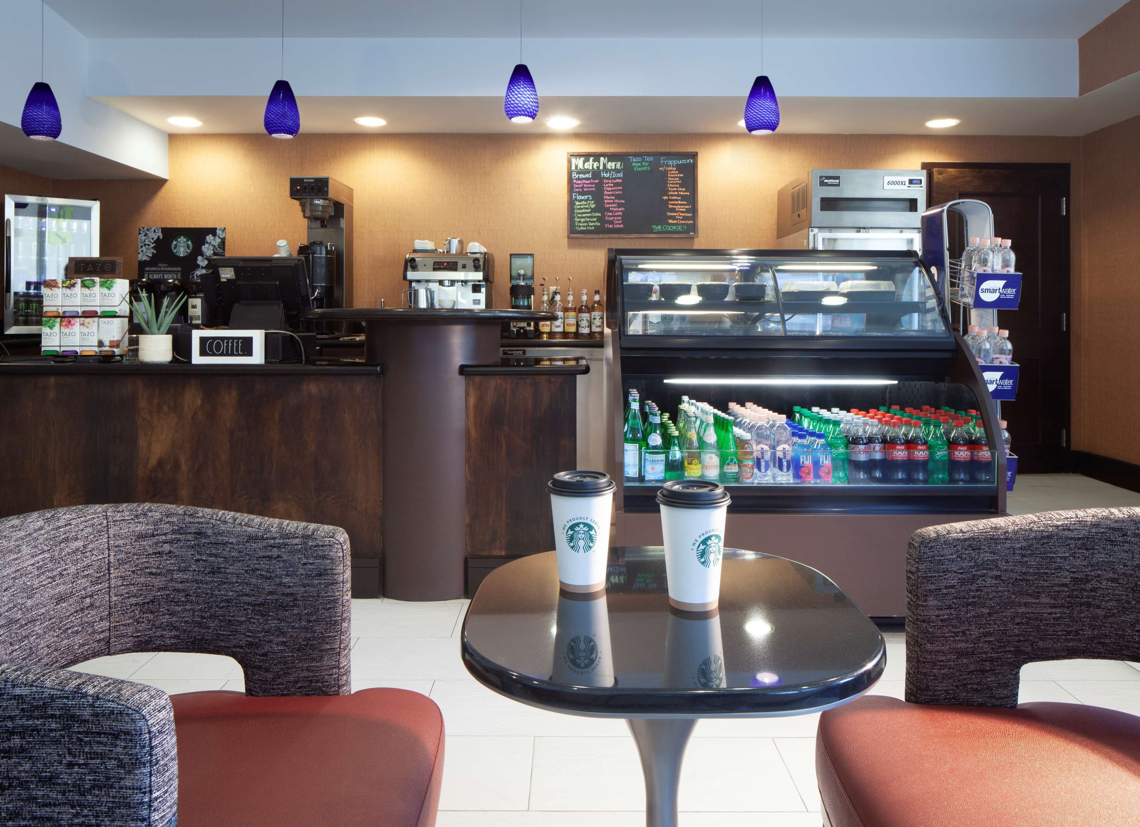 L’intérieur d’un café présente un comptoir, une machine à expresso et un présentoir contenant des boissons en bouteille. On y voit deux chaises et une table avec deux gobelets Starbucks.
