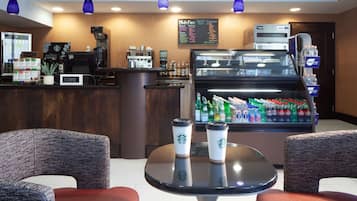 L’intérieur d’un café présente un comptoir, une machine à expresso et un présentoir contenant des boissons en bouteille. On y voit deux chaises et une table avec deux gobelets Starbucks.