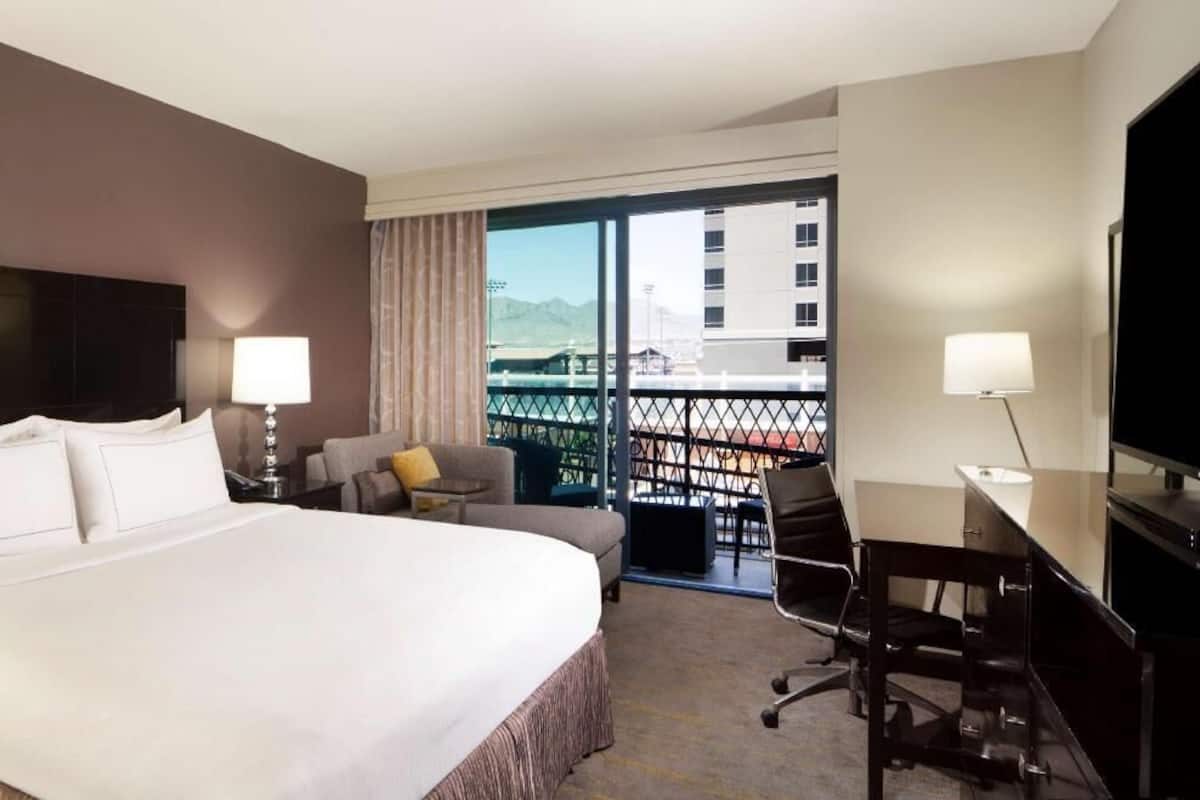 A hotel room with a bed, a desk, a chair, a TV, a balcony with a view of mountains, and a window with curtains.