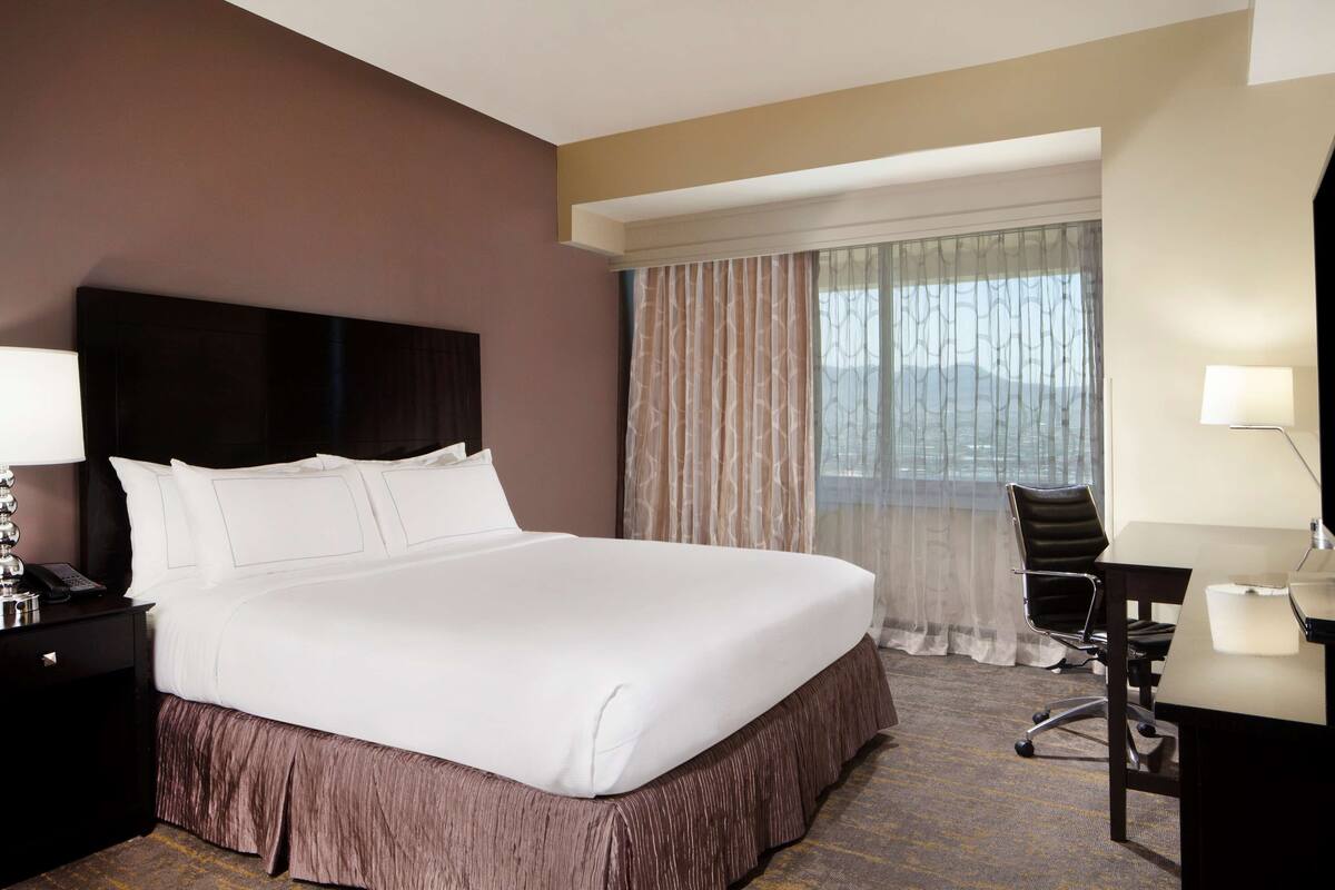 A hotel room with a large bed, a desk, a chair, a TV, and a window with curtains.