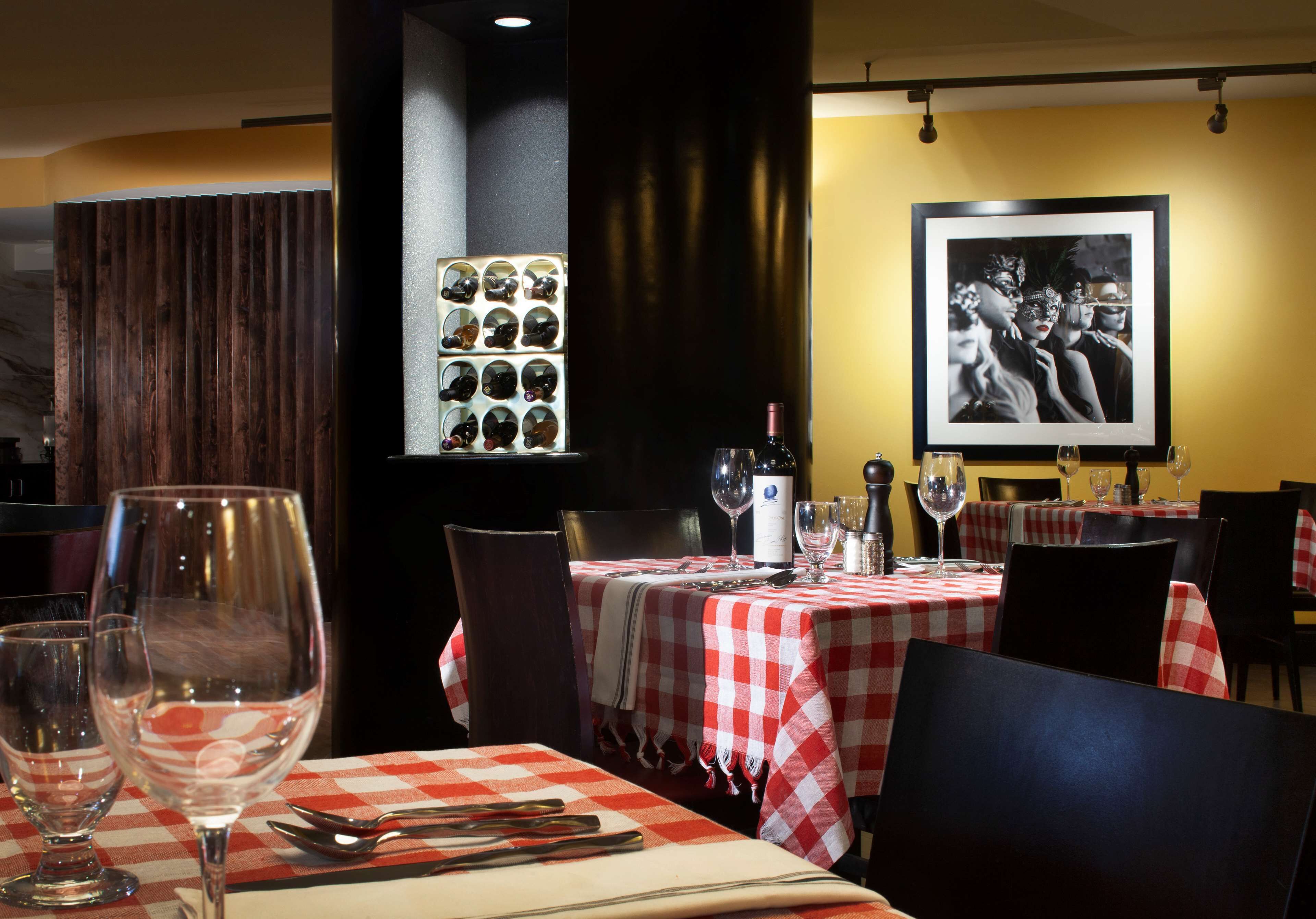 A restaurant interior with checkered tablecloths, wine glasses, and a wall-mounted display of wine bottles.