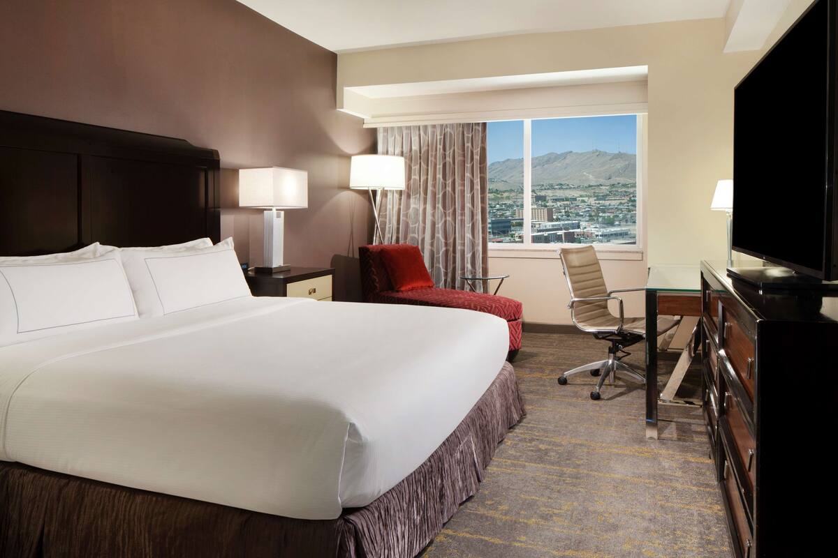 A hotel room with a large bed, a desk with a chair, a TV, and a window with a view of a city and mountains.