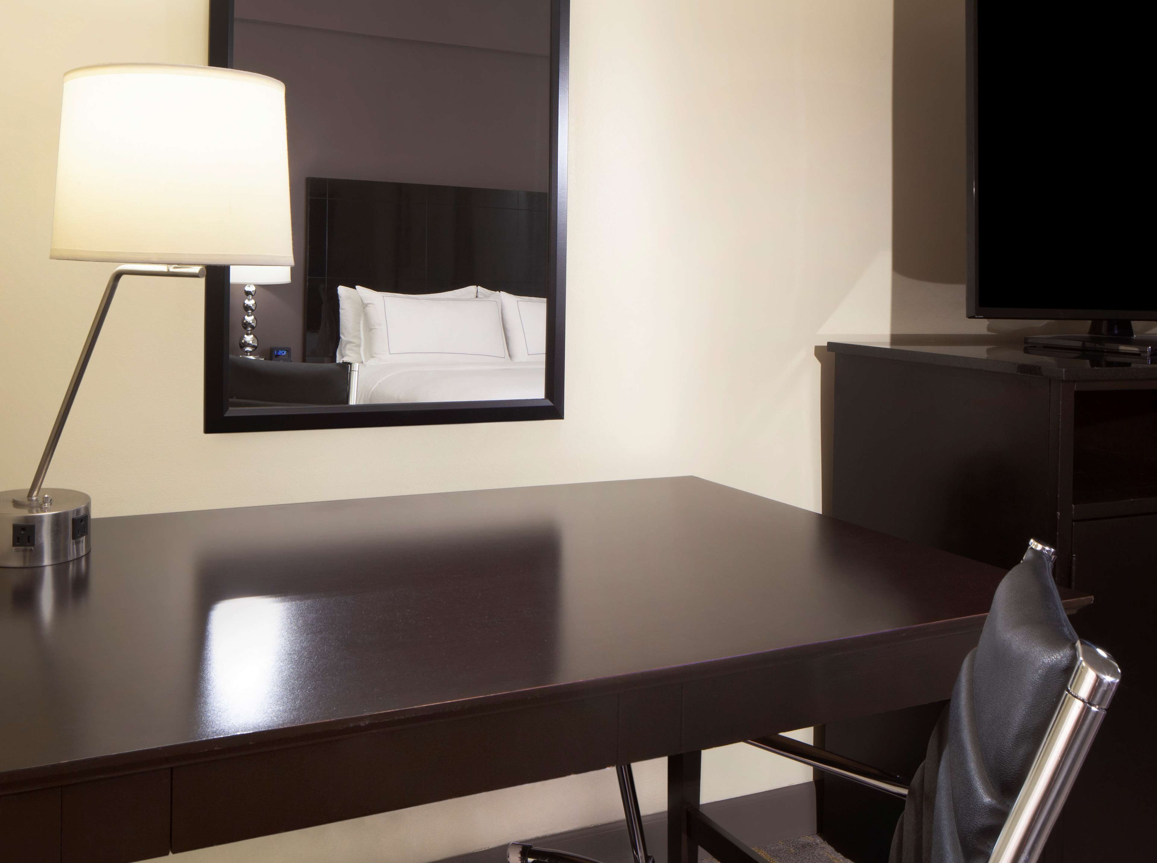 Habitación de hotel con un escritorio de madera oscura, una lámpara, una silla, un televisor y un espejo grande que refleja una cama.