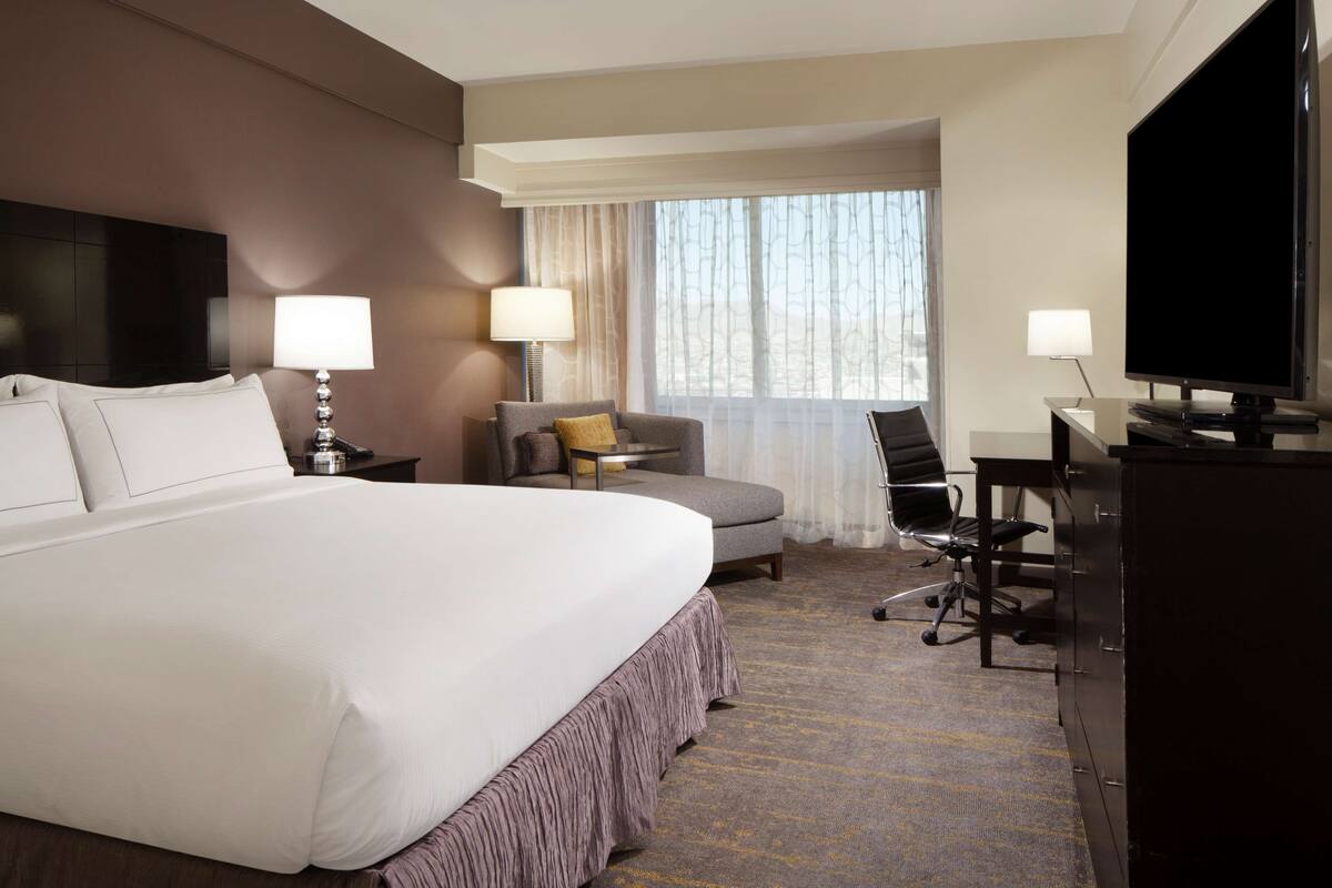 A hotel room with a large bed, a desk with a chair, a television, and a window with curtains.