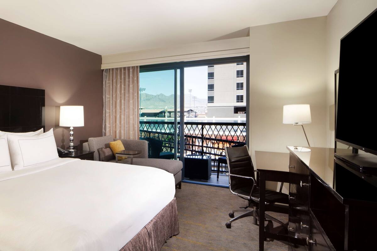 A hotel room with a large bed, a desk with a chair, a TV, and a balcony with a view of the city and mountains.