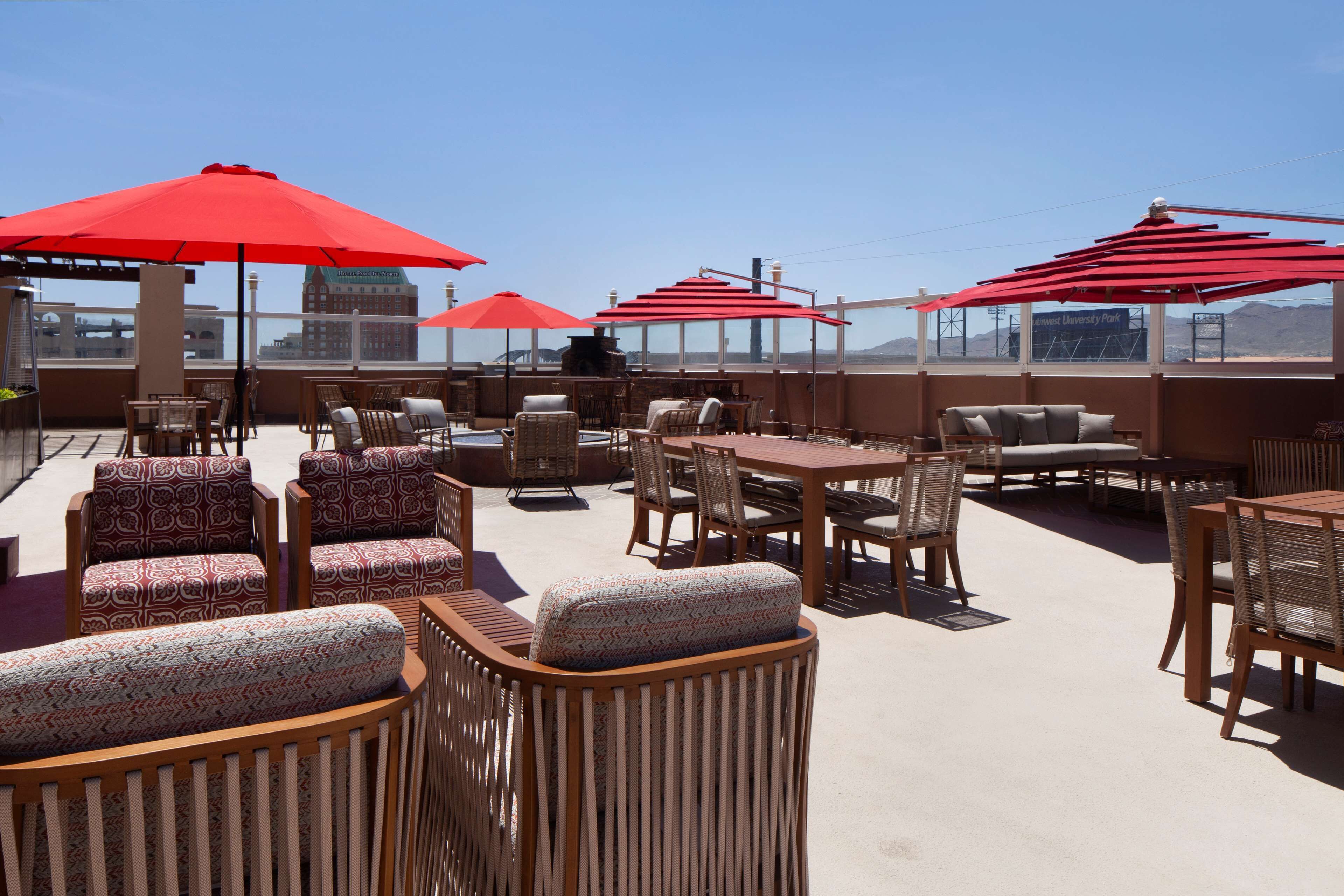 A rooftop terrace with outdoor seating, red umbrellas, and a view of a distant building.