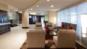 A spacious lobby with a reception desk, seating area with armchairs and sofas, a large window with sheer curtains, and a modern interior design.