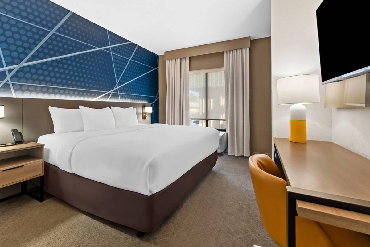 Habitación de hotel con una cama grande, mesitas de noche, un escritorio, una silla, un televisor y una ventana con cortinas.