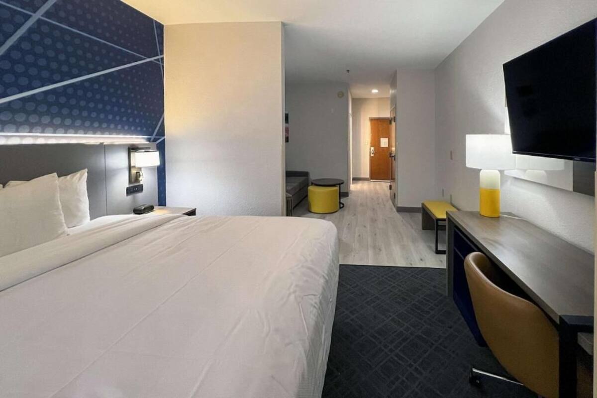 Una habitación de hotel con una cama grande, un escritorio con silla, un televisor y una mesita de noche con lámpara.