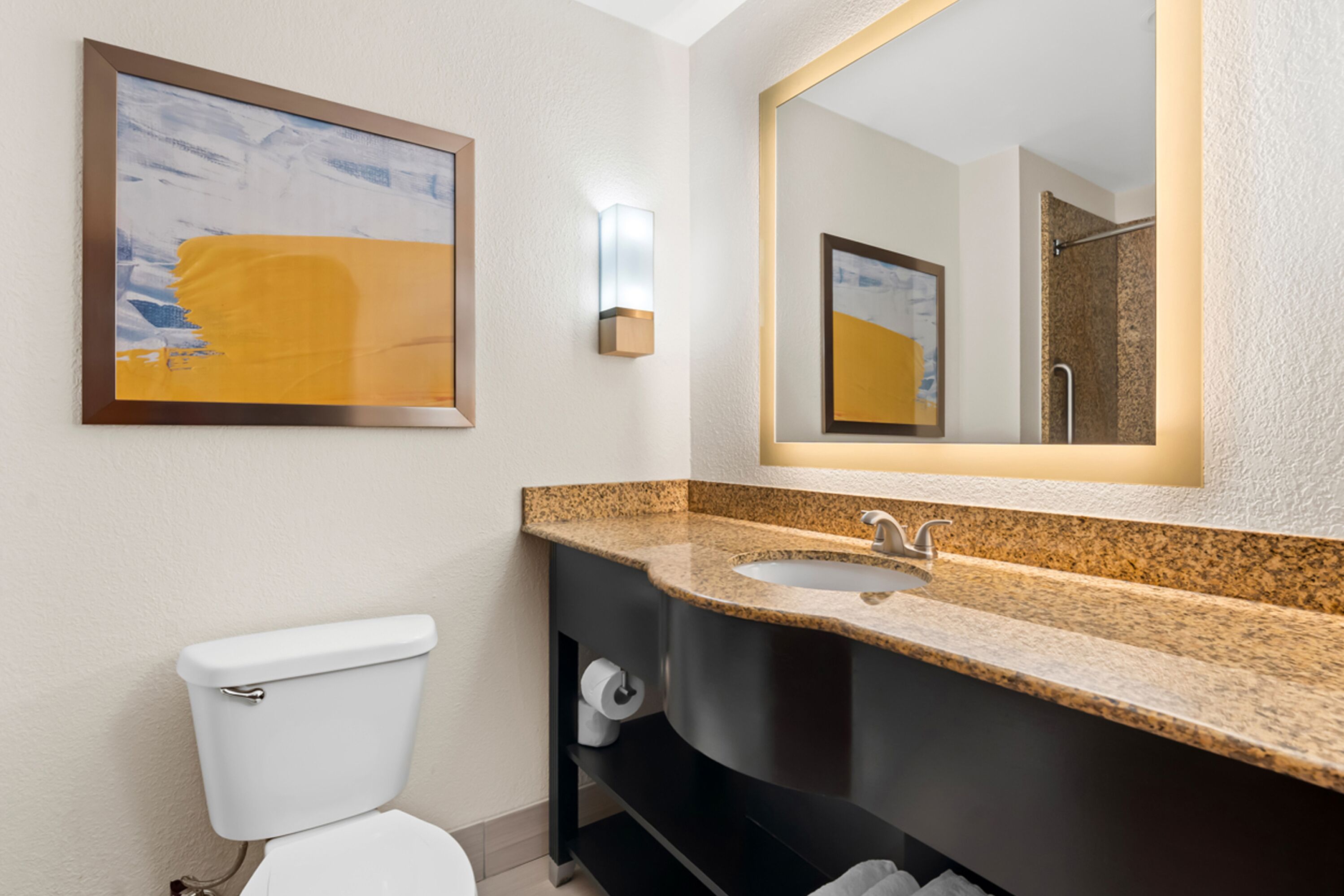 A modern bathroom with a white toilet, granite countertop sink, and a large mirror.