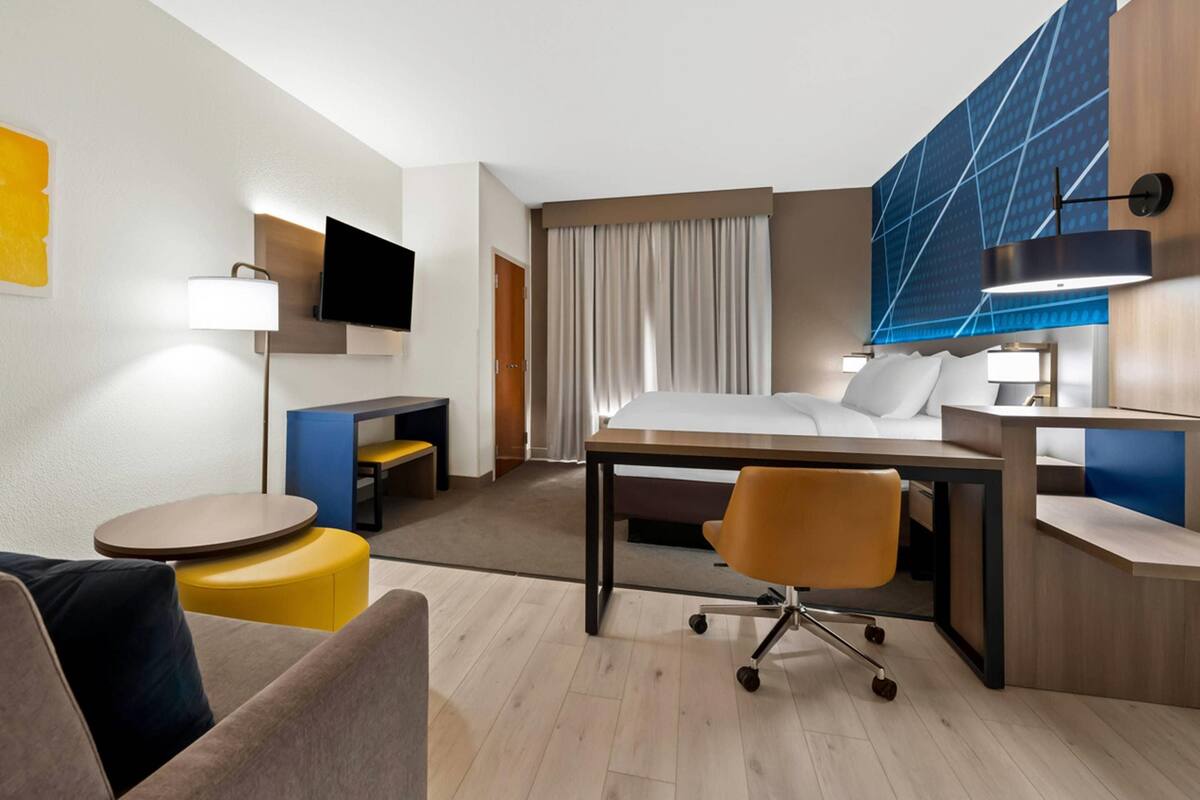 Habitación de hotel con cama, escritorio, silla, televisor y un ventanal con cortinas.