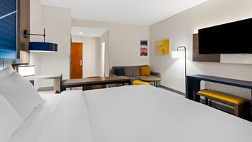 A modern hotel room with a large bed, a desk, a sofa, a small table, and a TV mounted on the wall.